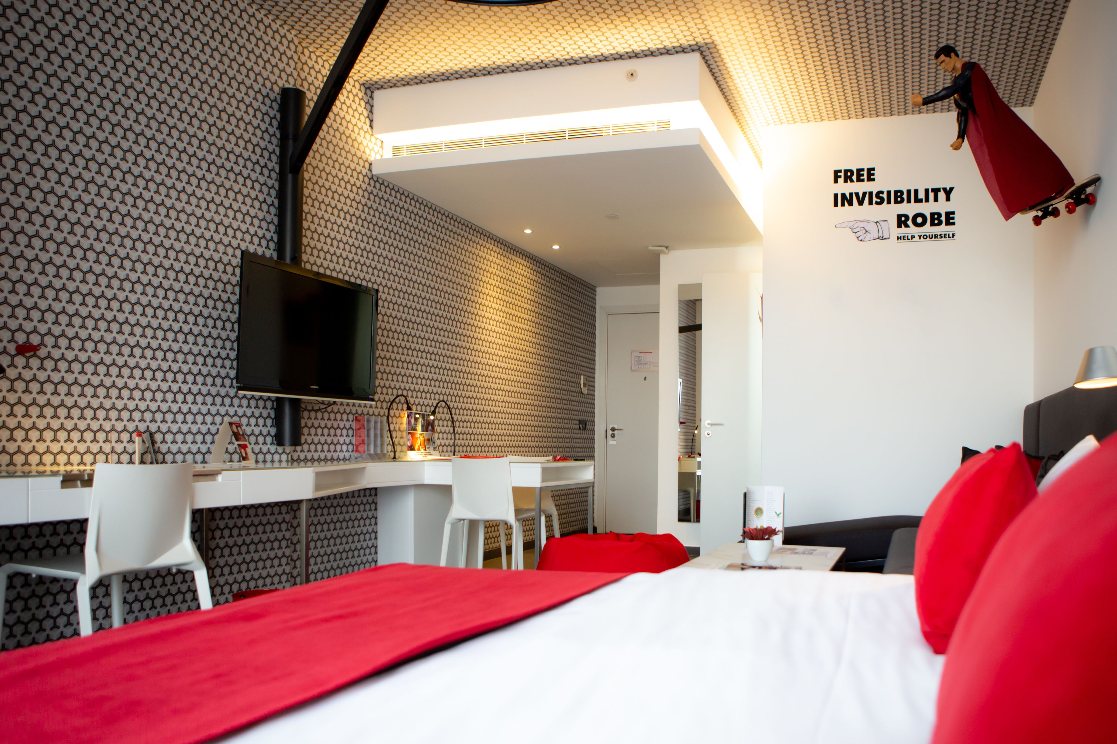 A modern hotel room with a white bed, red accents, a desk with a chair, a TV, and a wall with a superhero-themed artwork.