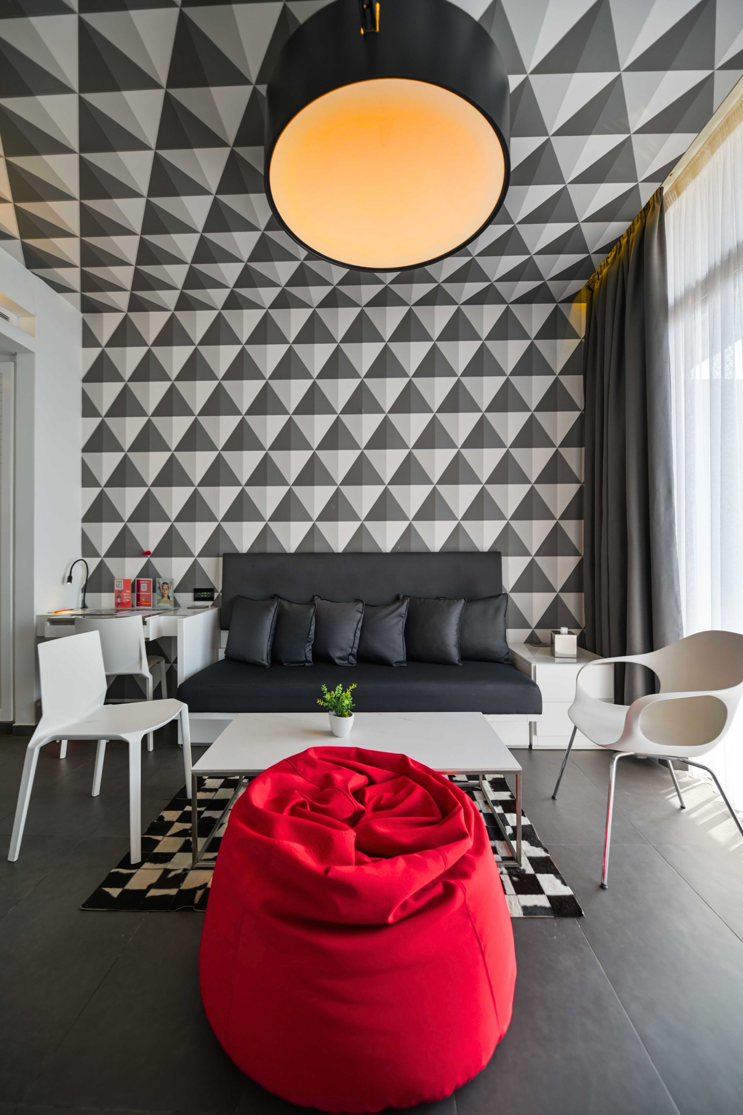 Un salon moderne avec un mur orné d’un motif géométrique, un canapé noir, des chaises blanches, un fauteuil en forme de poire rouge et une table basse centrale.