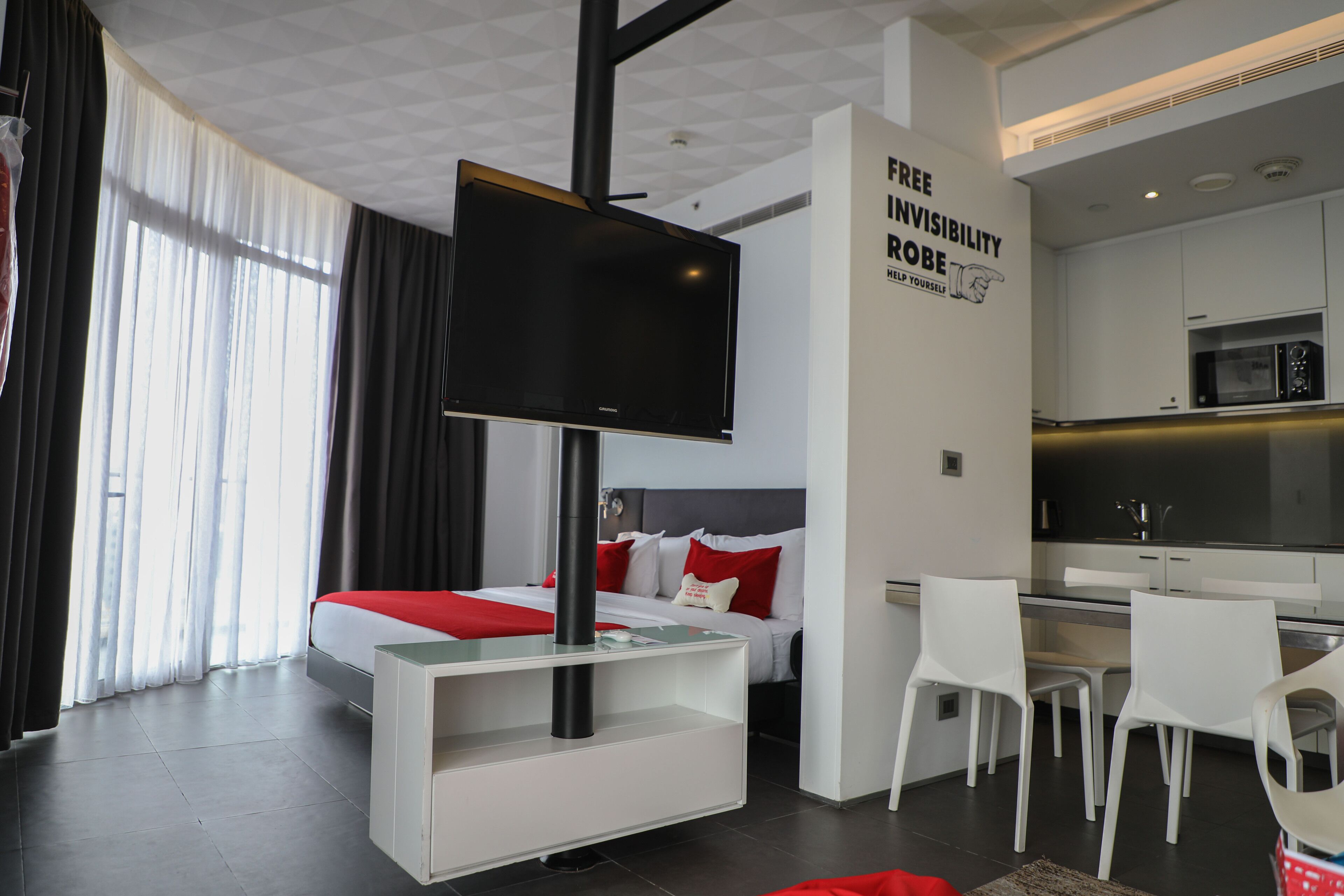 A modern hotel room with a bed, a TV mounted on a stand, a dining area with chairs, and a kitchen area.