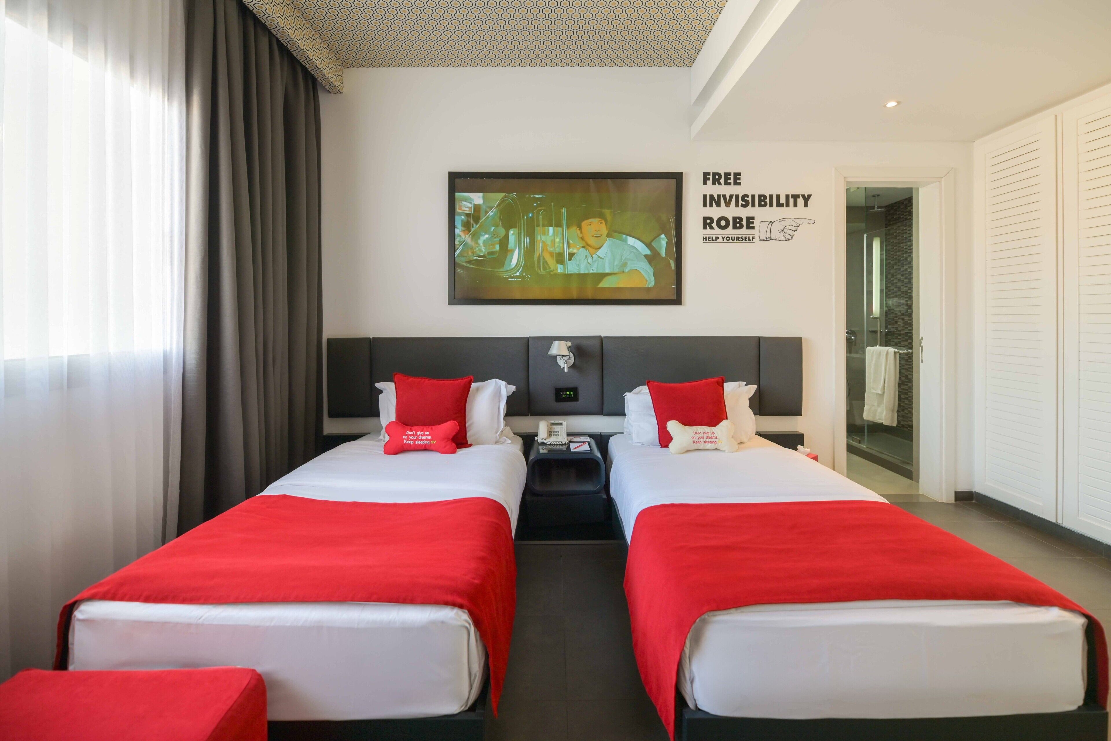 A hotel room with two beds, red bedding, a TV on the wall, and a motivational quote.