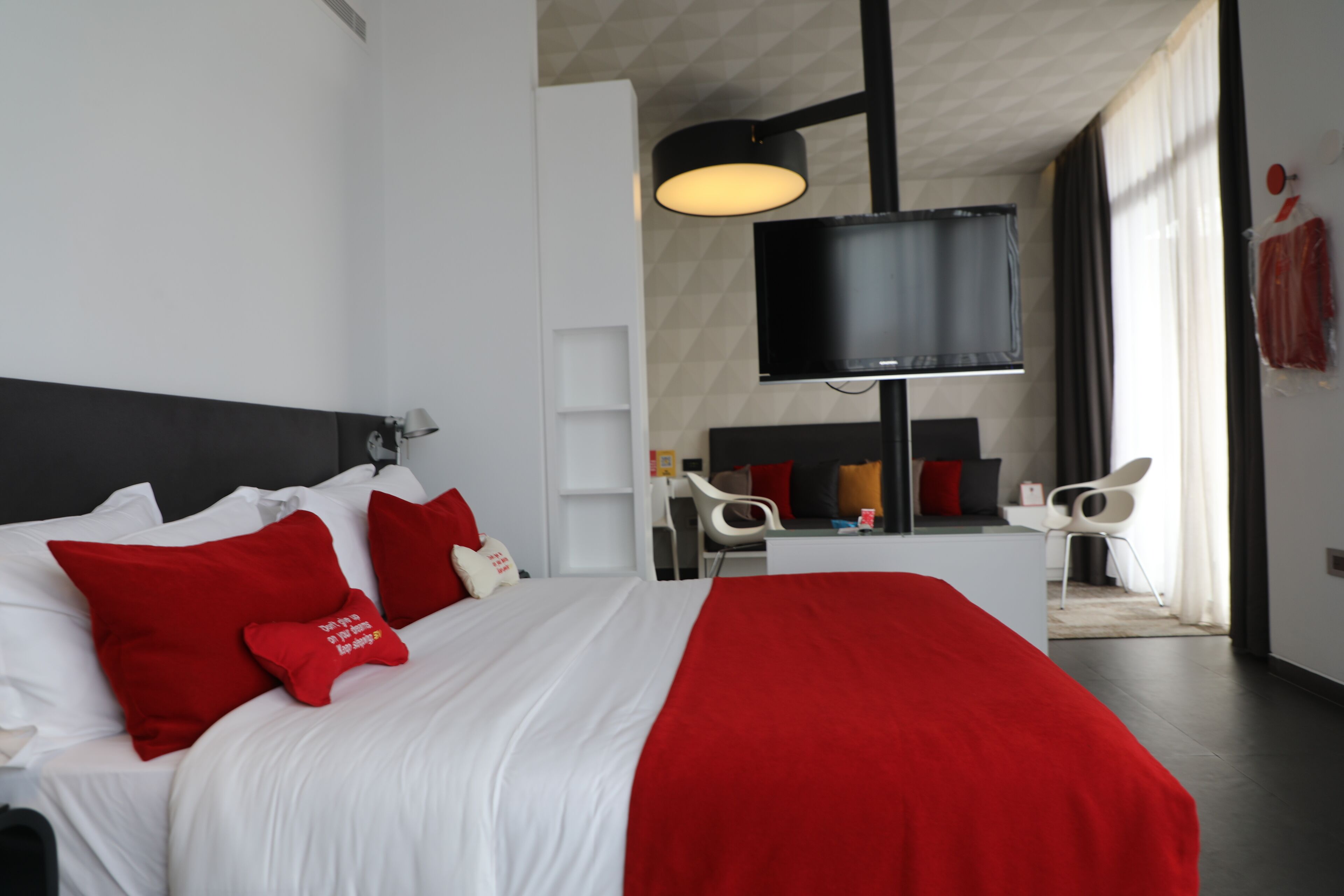 Una habitación de hotel moderna con una cama grande, ropa de cama roja y blanca, un televisor de pantalla plana y un área de descanso con un escritorio y sillas.