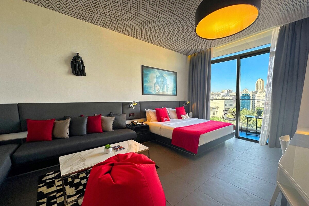 A modern hotel room with a large bed, a sofa, a coffee table, and a view of the city.