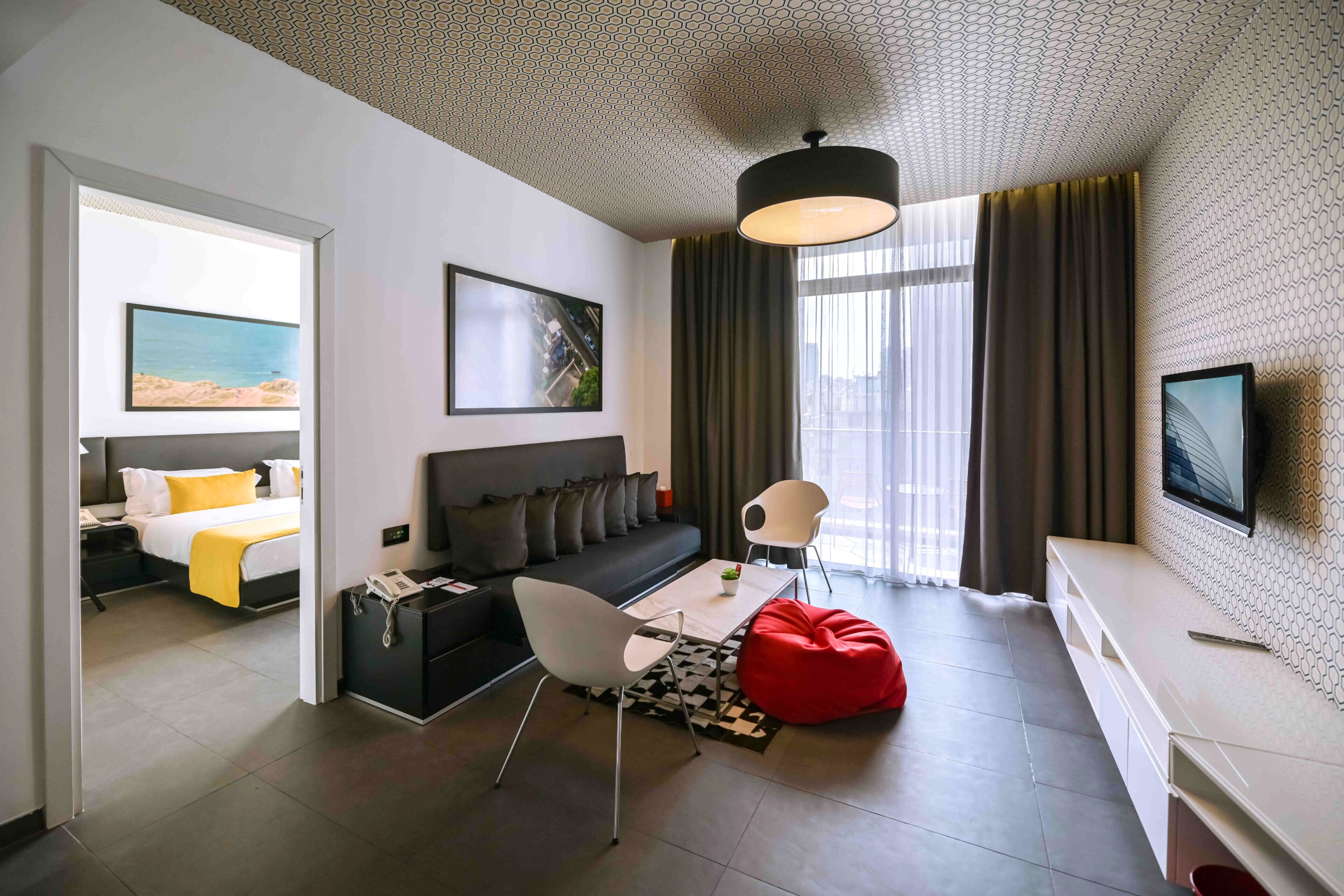 Una habitación de hotel moderna con un sofá, una mesa de comedor, un sillón de peluche rojo, un televisor y obras de arte en las paredes.