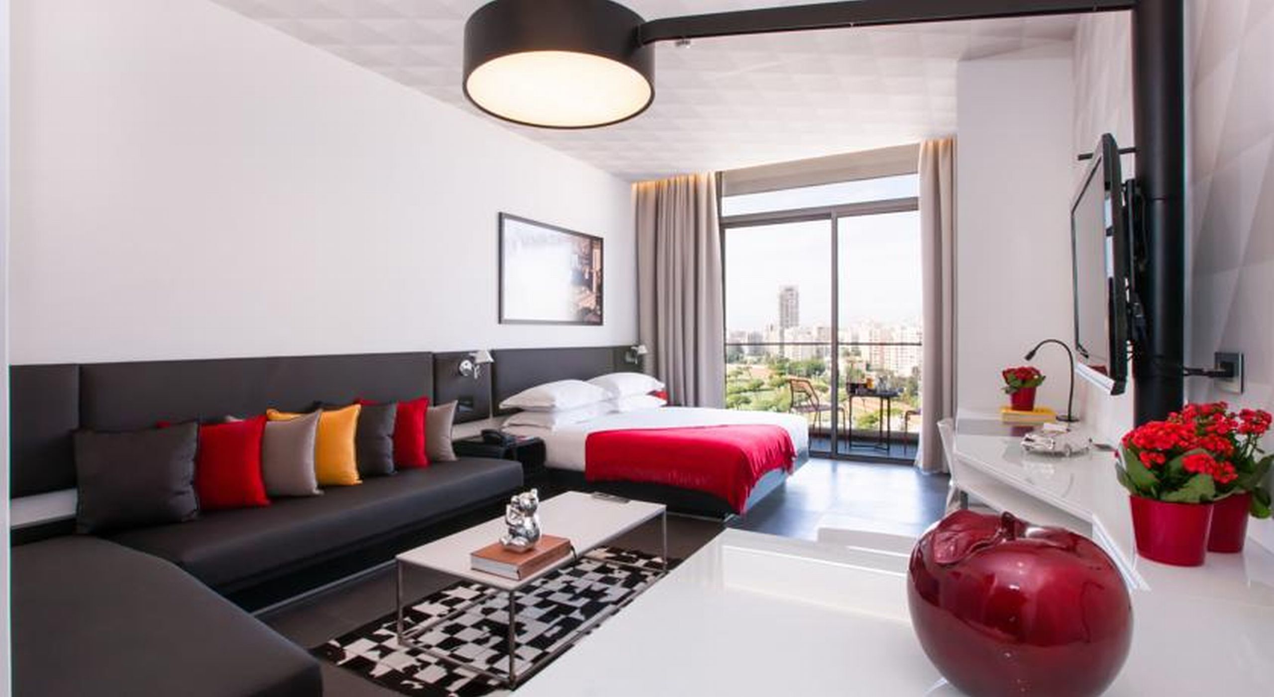 A modern hotel room with a large bed, a sofa, a coffee table, and a view of the city through large windows.