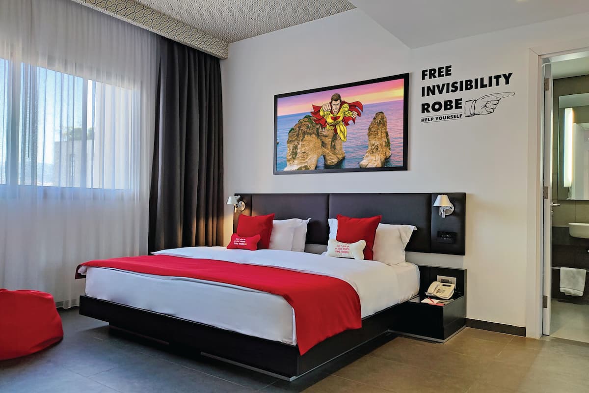 A modern hotel room with a large bed, a red and white color scheme, a framed picture on the wall, and a bathroom visible through an open door.