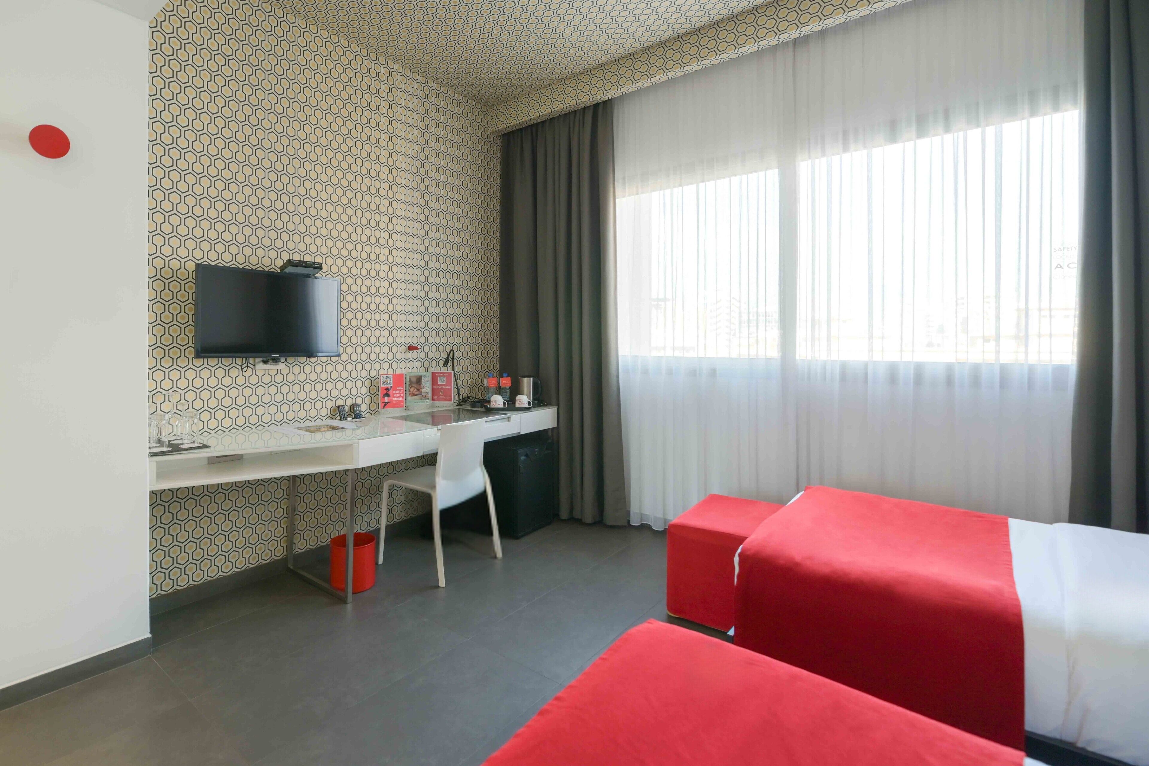 Quarto de hotel com escrivaninha, televisão, cama e janela com cortinas.