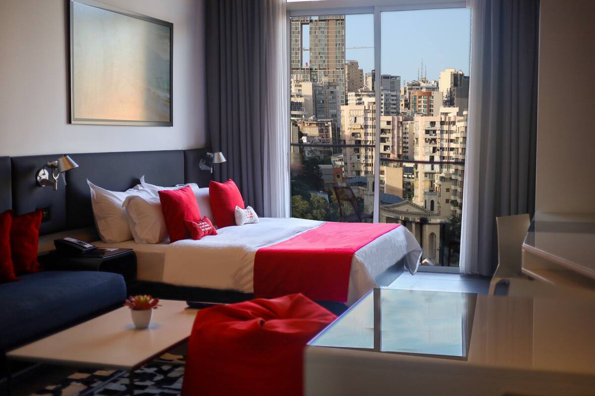 A hotel room with a large bed, a sofa, a small table with a plant, and a city view through the window.