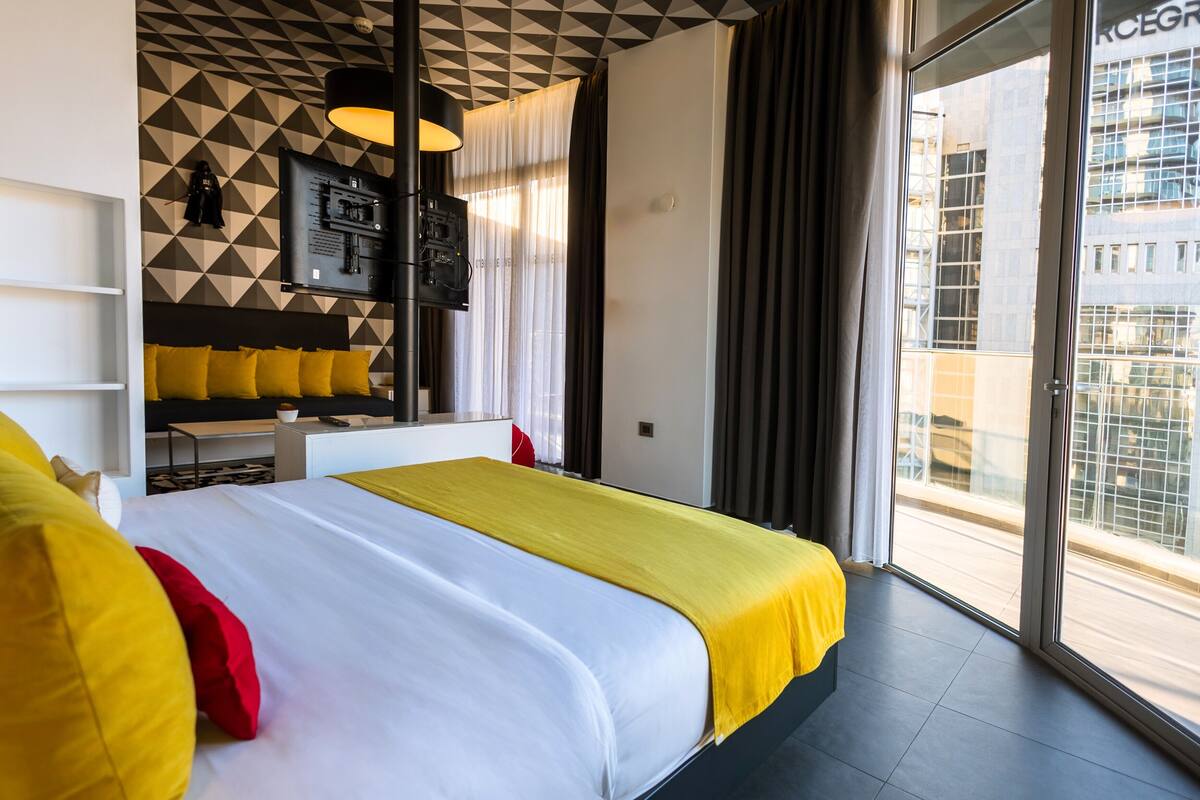 A modern hotel room with a large bed, a TV, a desk, and a view of the city.