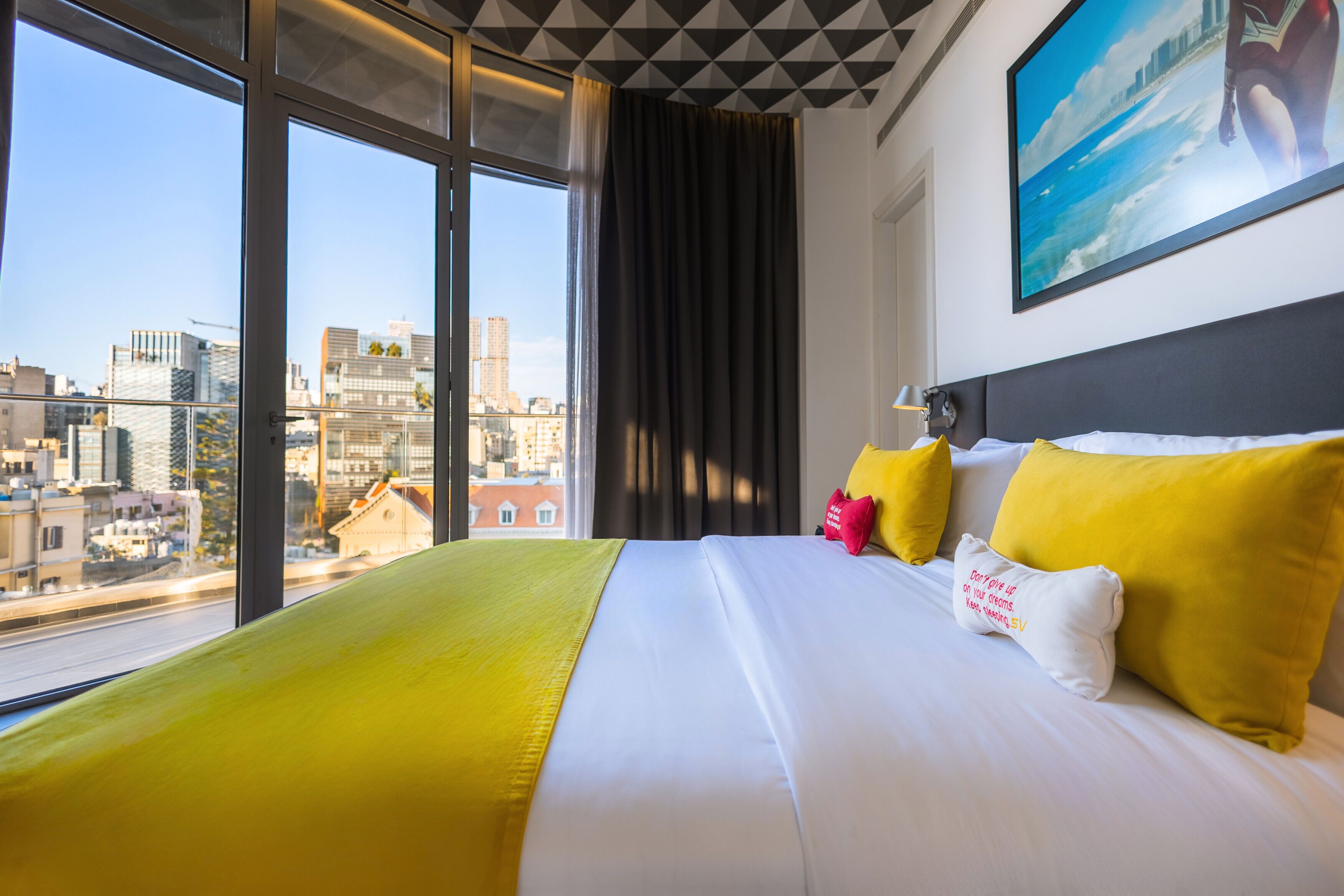 Hotellihuone, jossa on suuri sänky, keltaiset ja valkoiset vuodevaatteet, näkymä kaupunkiin suurista ikkunoista sekä taulu seinällä.