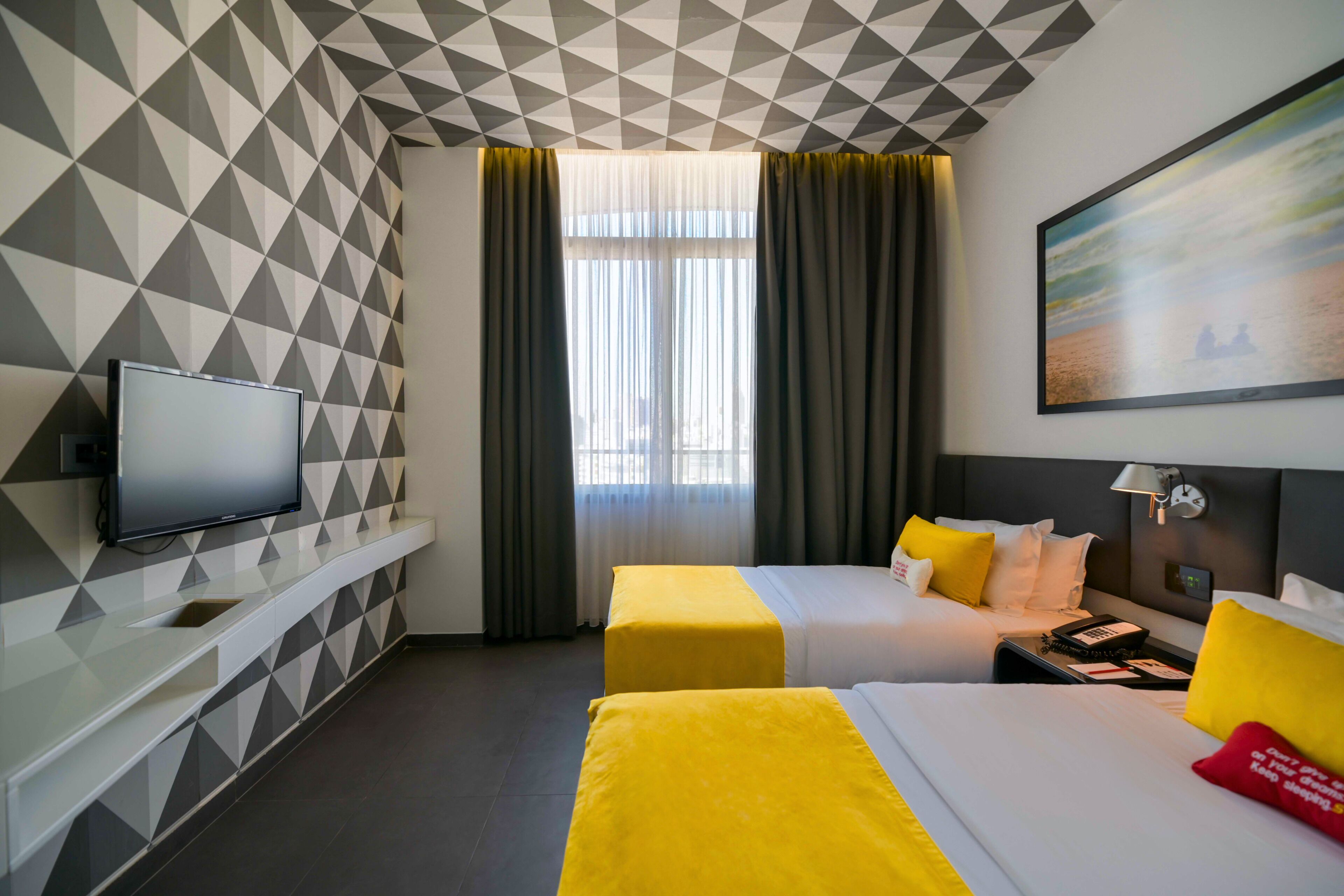 A hotel room with a geometric patterned wall, a flat-screen TV, two beds with yellow and white bedding, a window with curtains, and a painting on the wall.