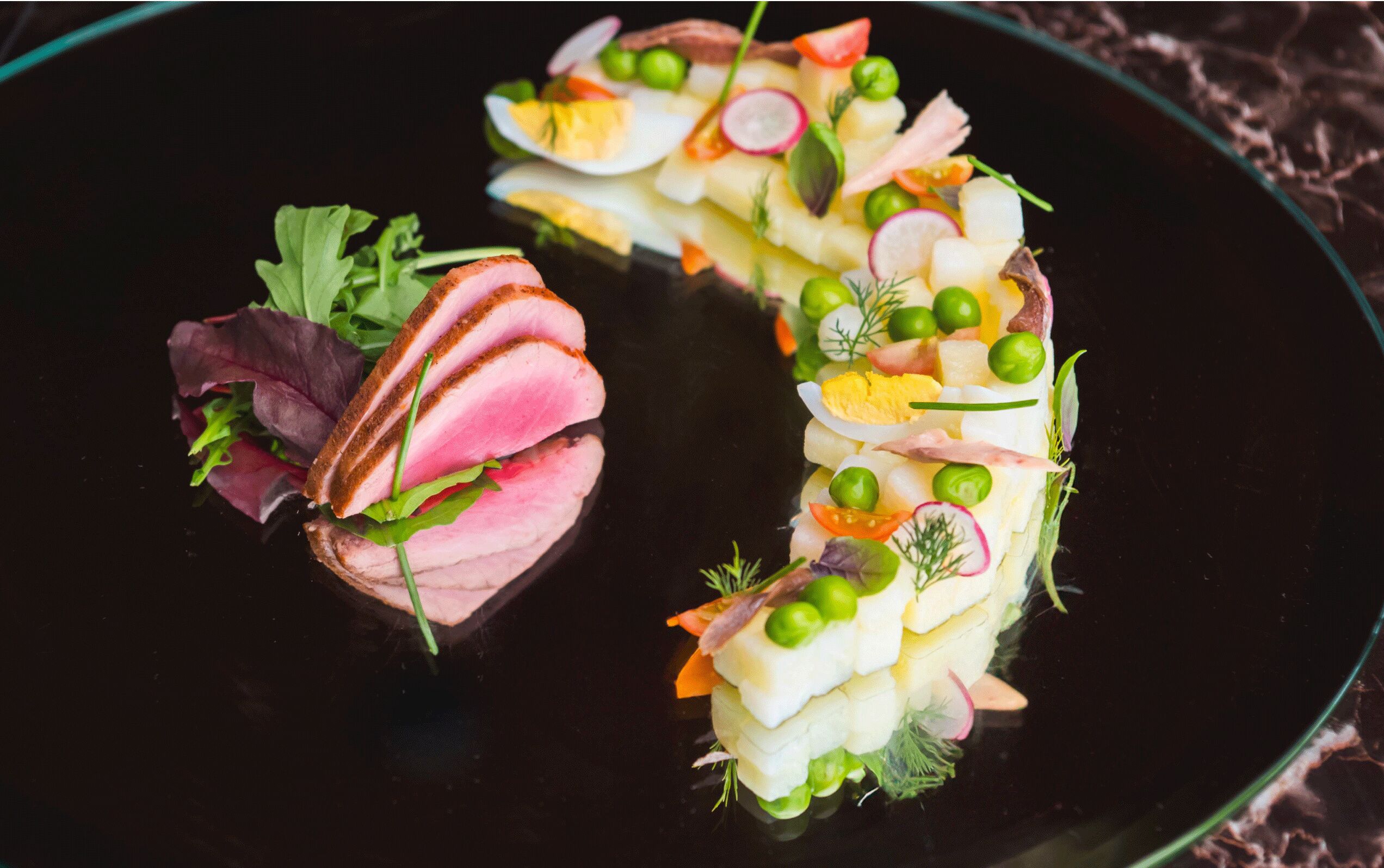 Un plato negro con dos preparaciones gourmet, una con centro rosado y guarnición verde, y otra con ingredientes blancos y verdes.