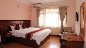 A hotel room with a brick headboard bed, white bedding, a wooden nightstand with a phone, a wooden dresser with a mirror, and a window with sheer curtains. - Hotel Brihaspati (Kathmandu)