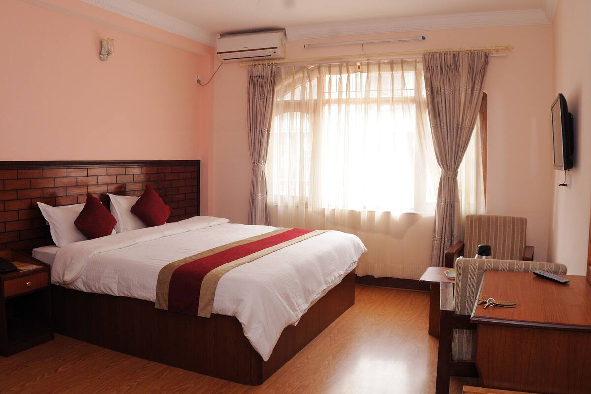 A hotel room with a brick headboard bed, white bedding, a wooden nightstand with a phone, a wooden dresser with a mirror, and a window with sheer curtains.