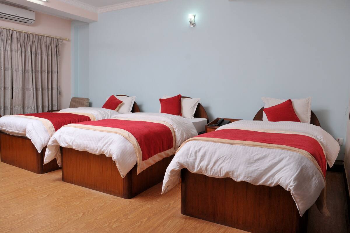 A hotel room with three wooden beds, each with white and red bedding. There is a window with curtains, a wall-mounted air conditioner, and a bedside table with a lamp.