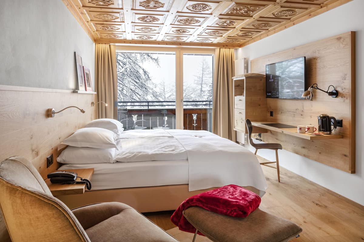 A modern hotel room with a wooden ceiling, a large window with a view of snow-covered trees, a bed with white linens, a bedside table with a lamp, a desk with a chair, and a mounted TV.