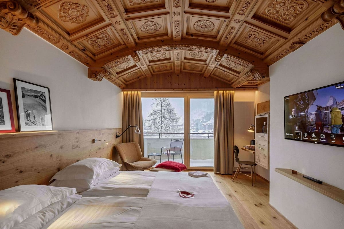 A hotel room with a wooden ceiling, a bed with white linens, a chair, a desk with a lamp, and a TV mounted on the wall.