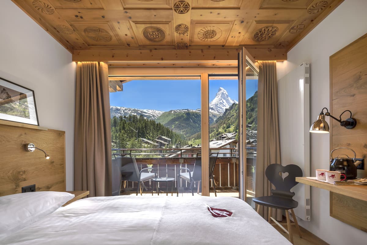 A hotel room with a bed, a desk with a kettle, a chair, and a balcony with a mountain view.