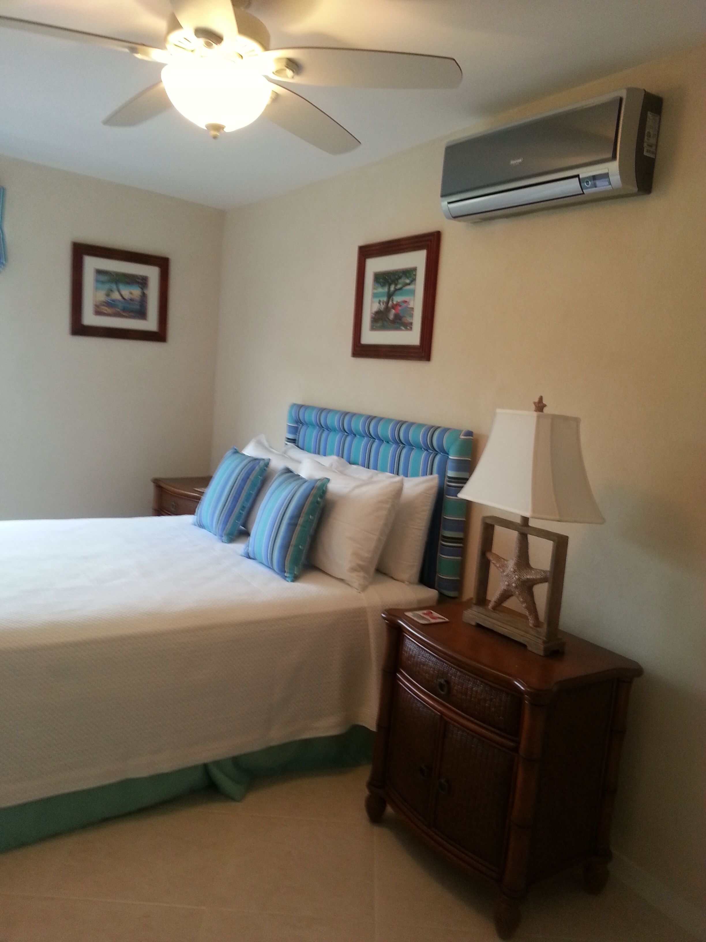 A bedroom with a bed, nightstands, a lamp, an air conditioner, and two framed pictures on the wall.