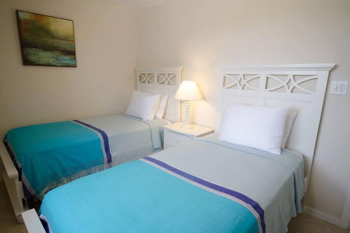 Una habitación con dos camas, cada una con cabecero blanco y colcha color turquesa. Entre las camas, una mesita de noche con una lámpara.