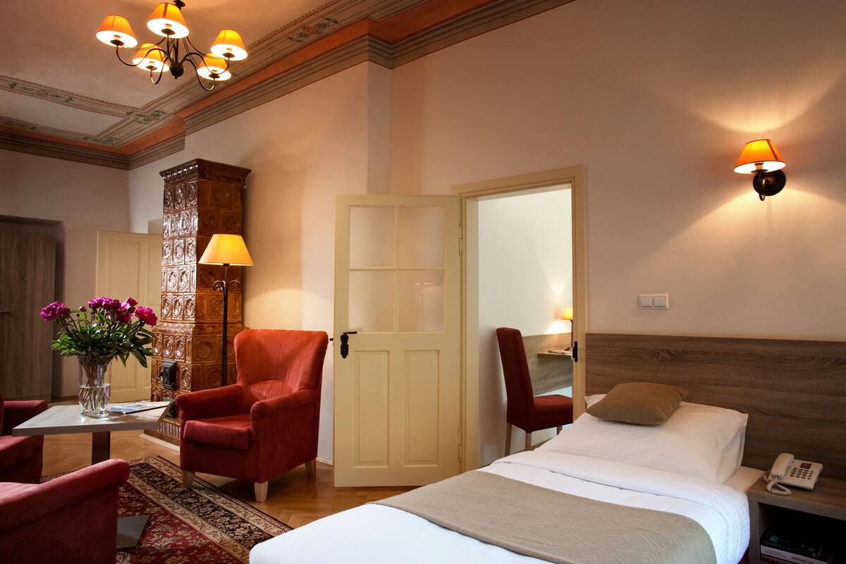 Habitación de hotel con una cama, dos sillones rojos, un aparador de madera, un jarrón con flores y una araña.