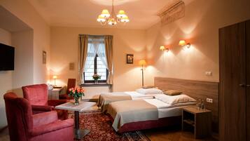 A hotel room with two beds, a red armchair, a small table with a vase of flowers, and a wooden wardrobe.
