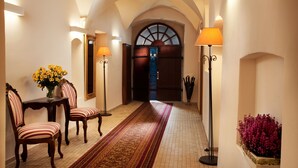 Un couloir avec un long tapis, deux fauteuils, une table ornée de fleurs et deux lampadaires.