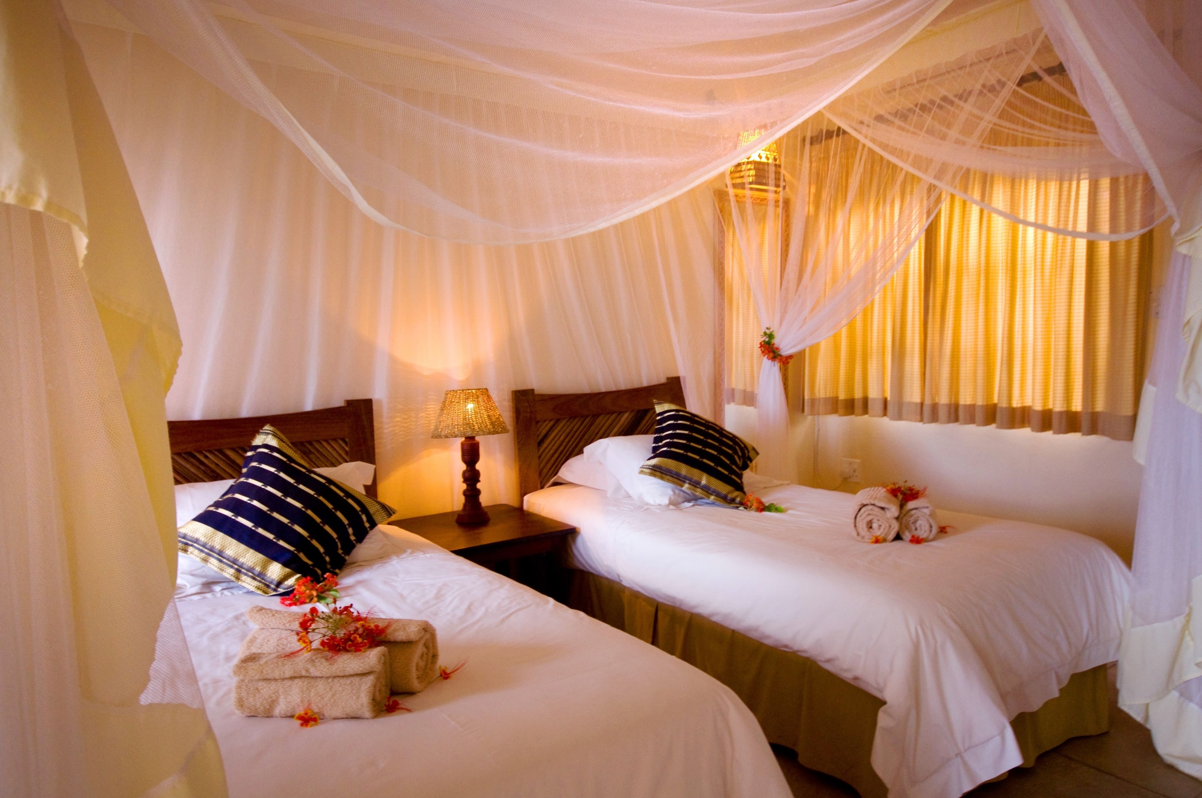 A hotel room with two beds, each with a pillow and a rolled towel. There is a nightstand with a lamp between the beds.