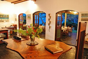 A wooden dining table with a vase of flowers, a chess set, and a book. There are chairs around the table, and a view of the outdoors through an arched doorway. - Casa Rex by Sol Resorts (Vilanculos)