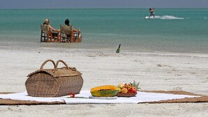 A beach picnic setup with a woven basket, fruits, and a table on the sand. - Casa Rex by Sol Resorts (Vilanculos)