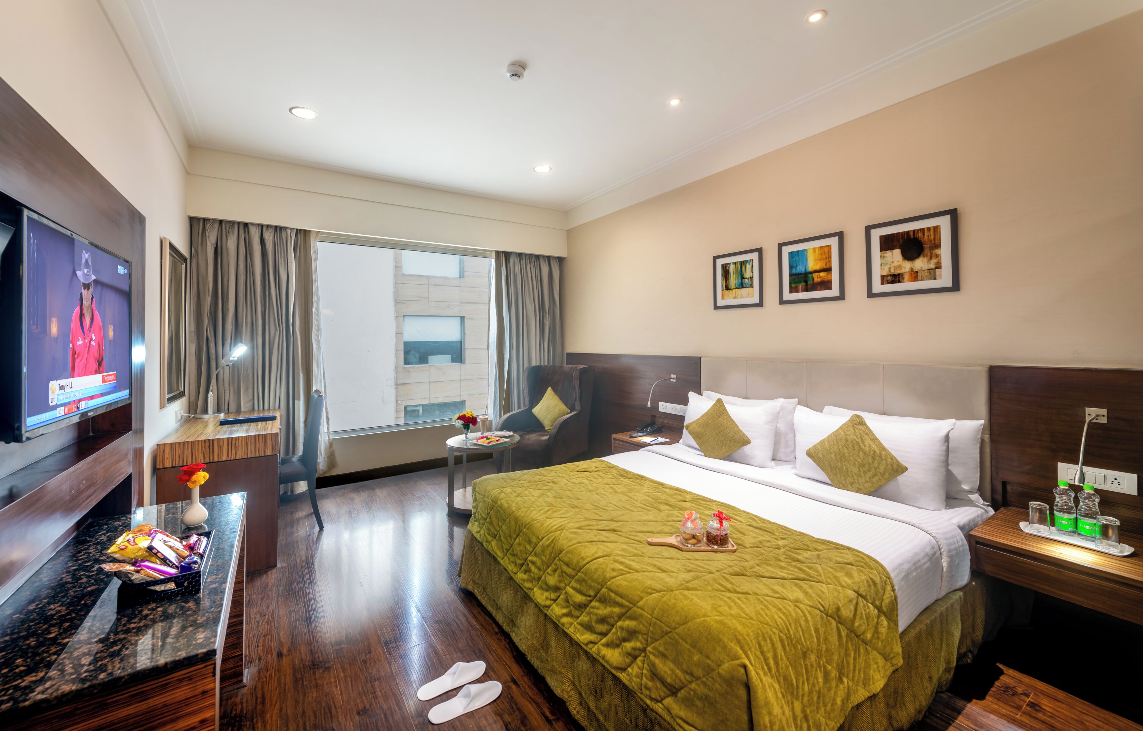A hotel room with a bed, a TV, a desk, a chair, a window, and artwork on the wall.