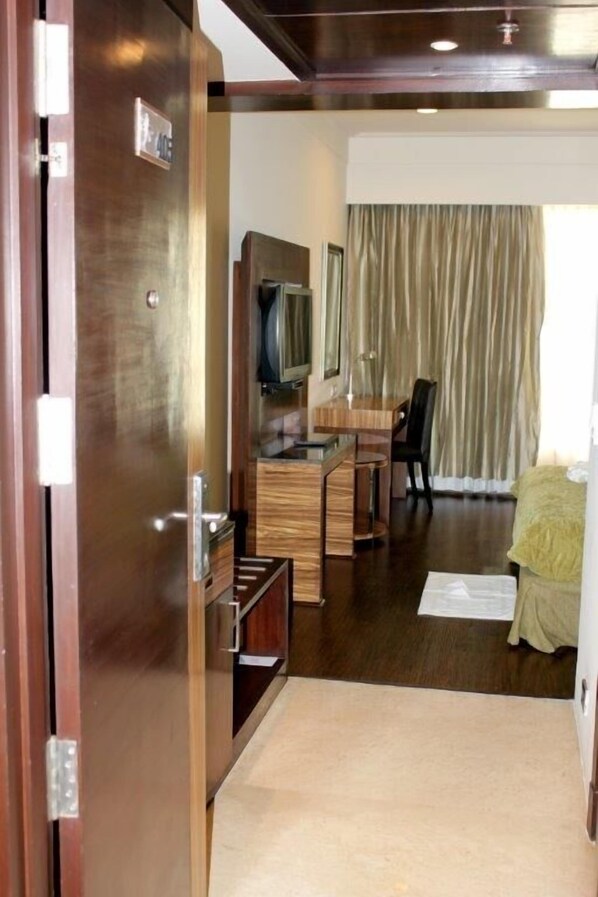A hotel room with a wooden door, a desk with a chair, a television, and a bed with a yellow blanket.