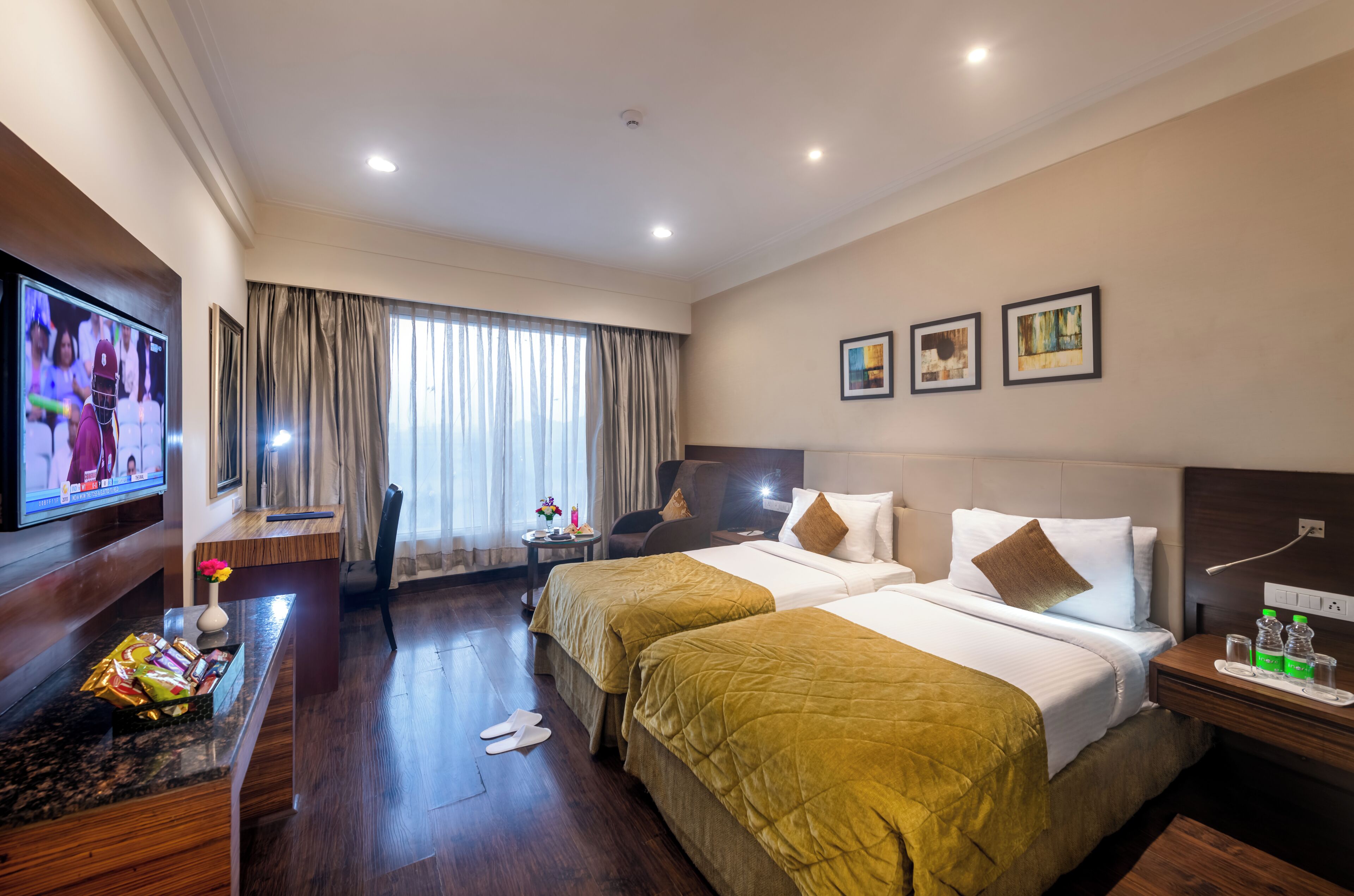 A hotel room with two beds, a TV, a desk with a vase, and a window with curtains.