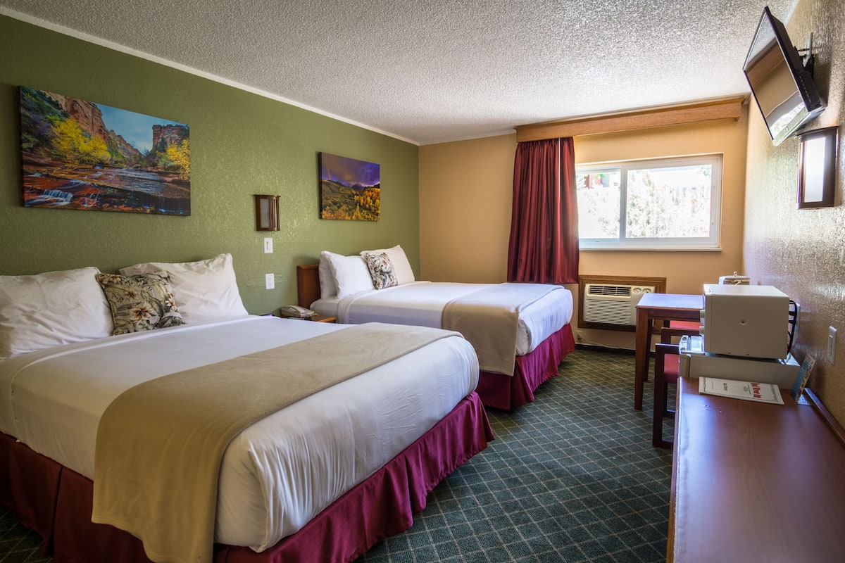 A hotel room with two beds, a desk, a window with curtains, and artwork on the walls.