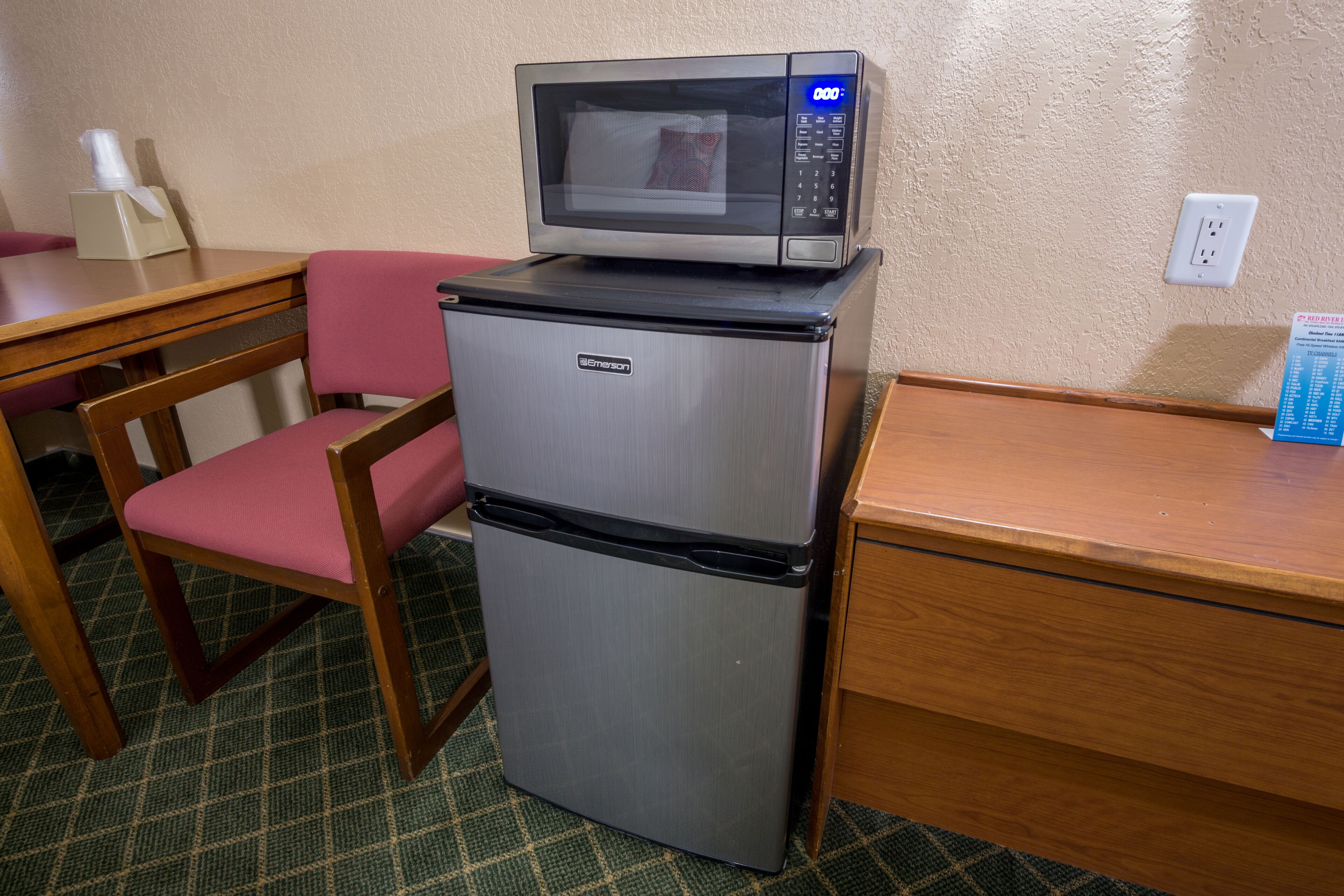 A compact kitchenette with a microwave on top of a refrigerator, a wooden table, a chair, and a tissue box.