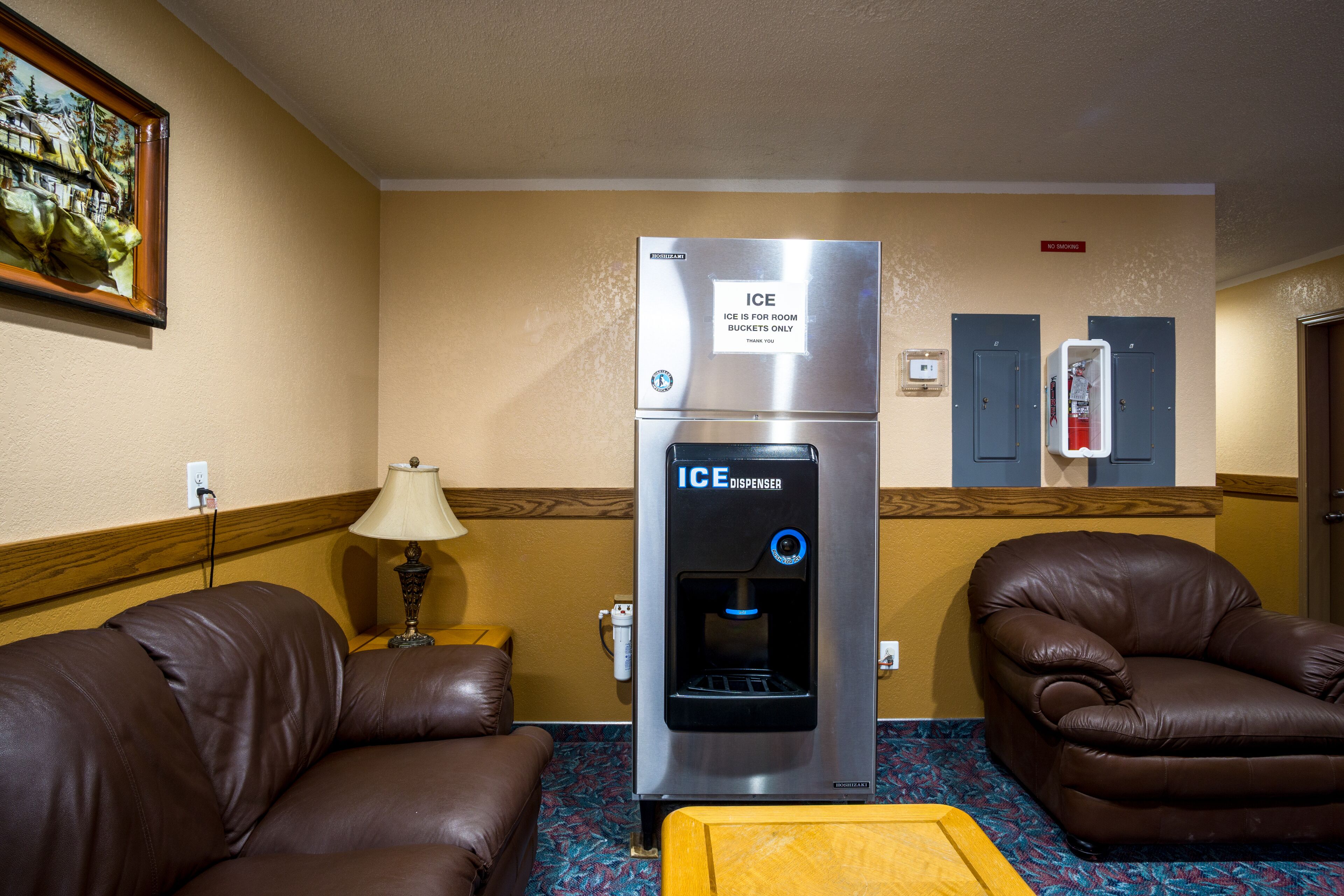 A hotel room with a minibar, a water dispenser, a couch, a chair, and a painting on the wall.