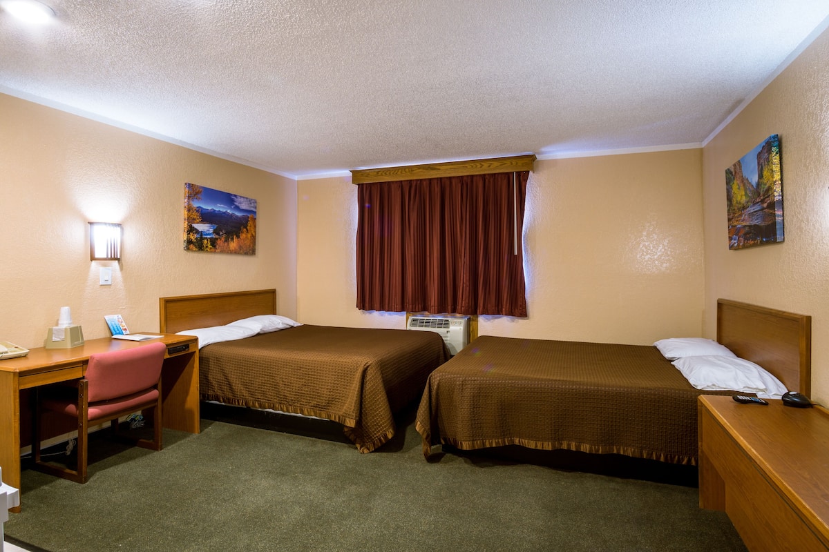 A hotel room with two beds, a desk, a chair, a lamp, and a painting on the wall.