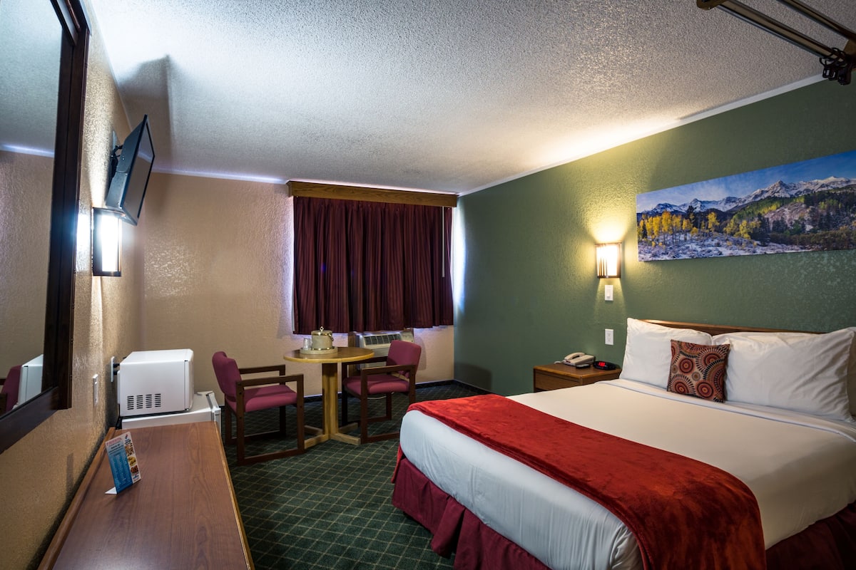A hotel room with a bed, a desk with a chair, a small table, a heater, a painting on the wall, and a window with curtains.
