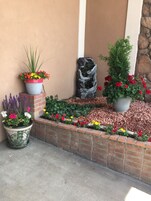 Um jardim ajardinado com diversos vasos de plantas e flores, uma escultura de pedra decorativa e uma borda de tijolos.