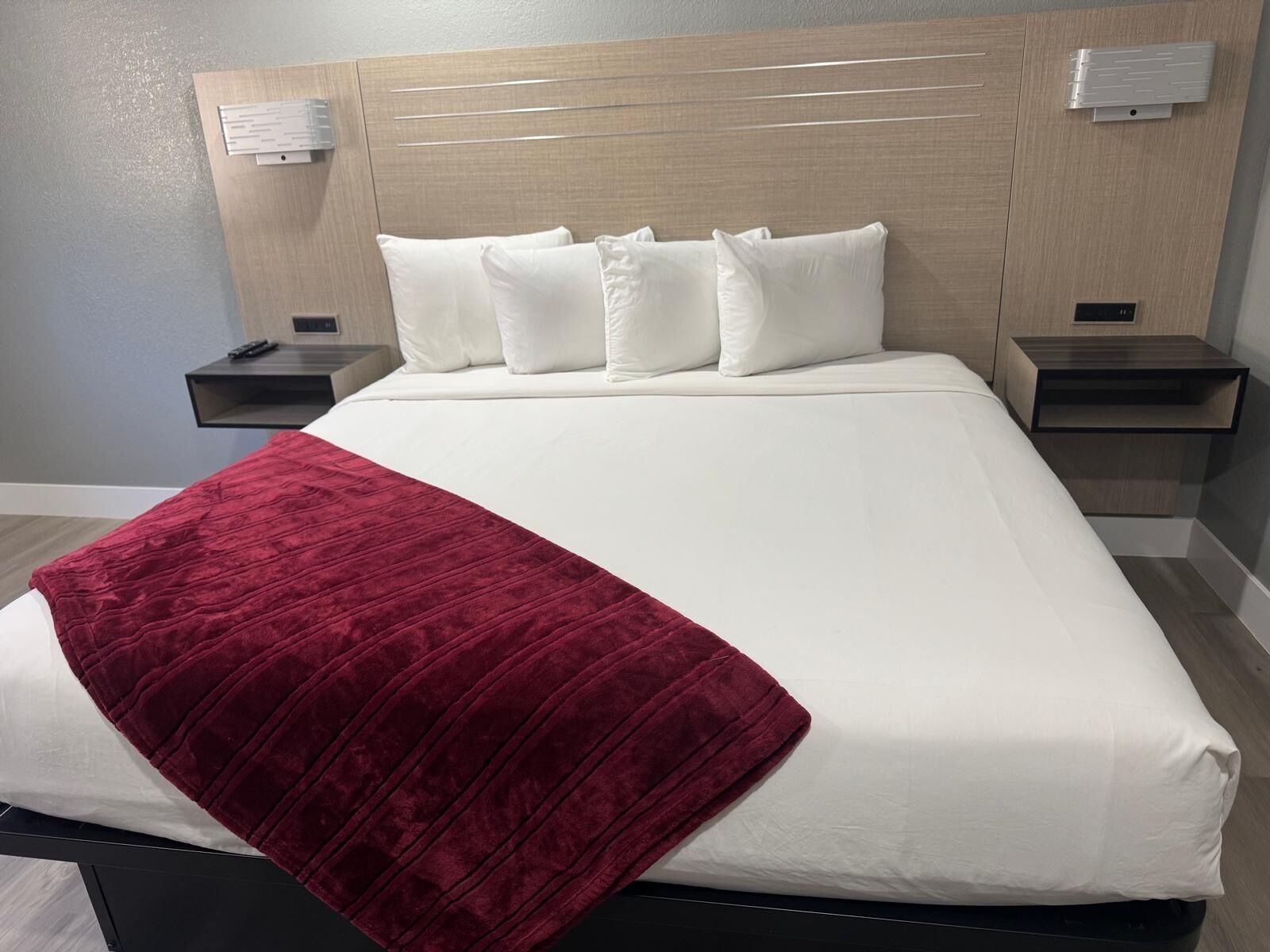 A hotel room with a large bed, white bedding, a red throw blanket, wooden headboard, bedside tables, and wall-mounted lights.