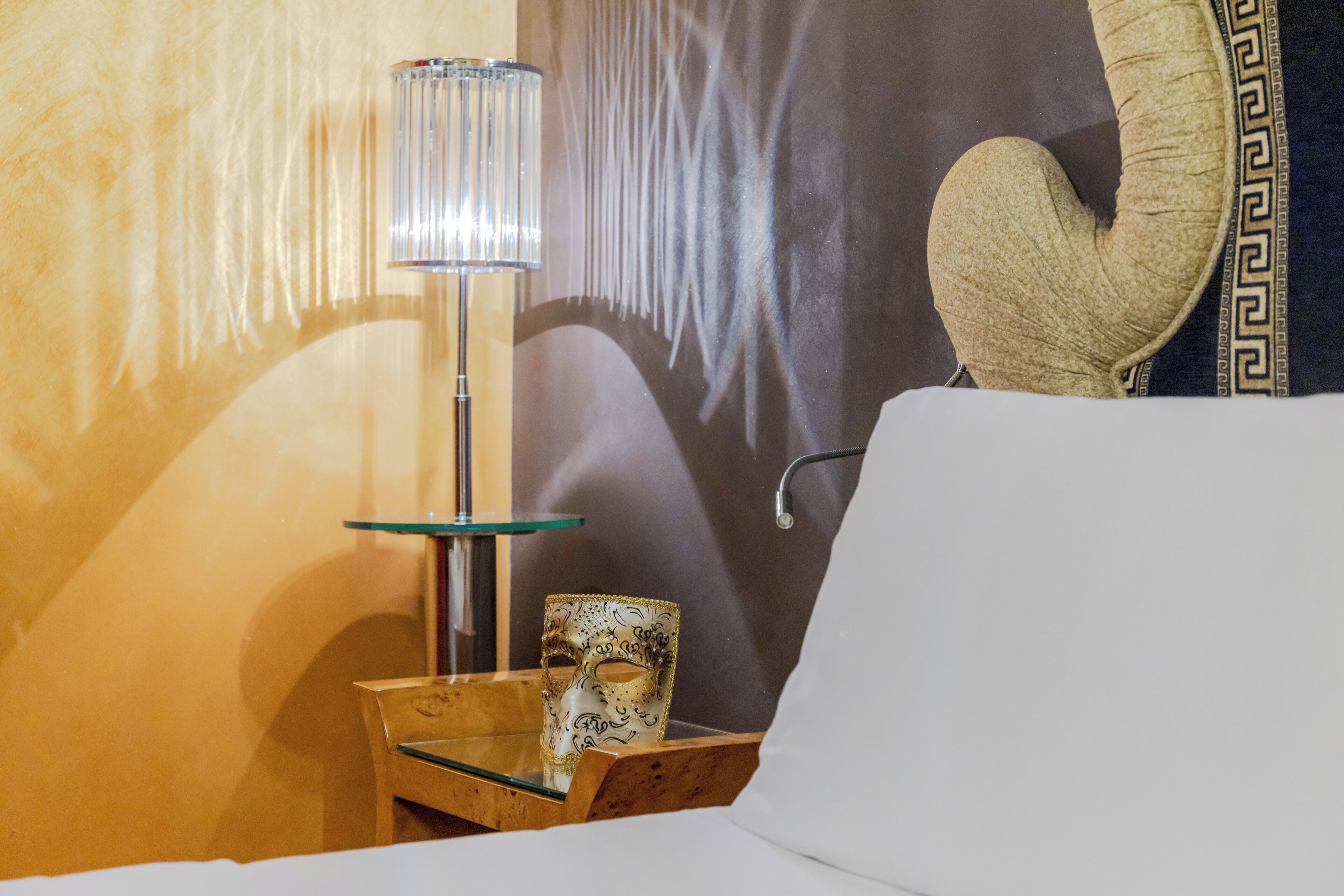 Een hotelkamer met een bed, een glazen bijzettafeltje met een lamp, een decoratief masker en een muur met een geometrisch patroon.