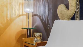 Een hotelkamer met een bed, een glazen bijzettafeltje met een lamp, een decoratief masker en een muur met een geometrisch patroon.