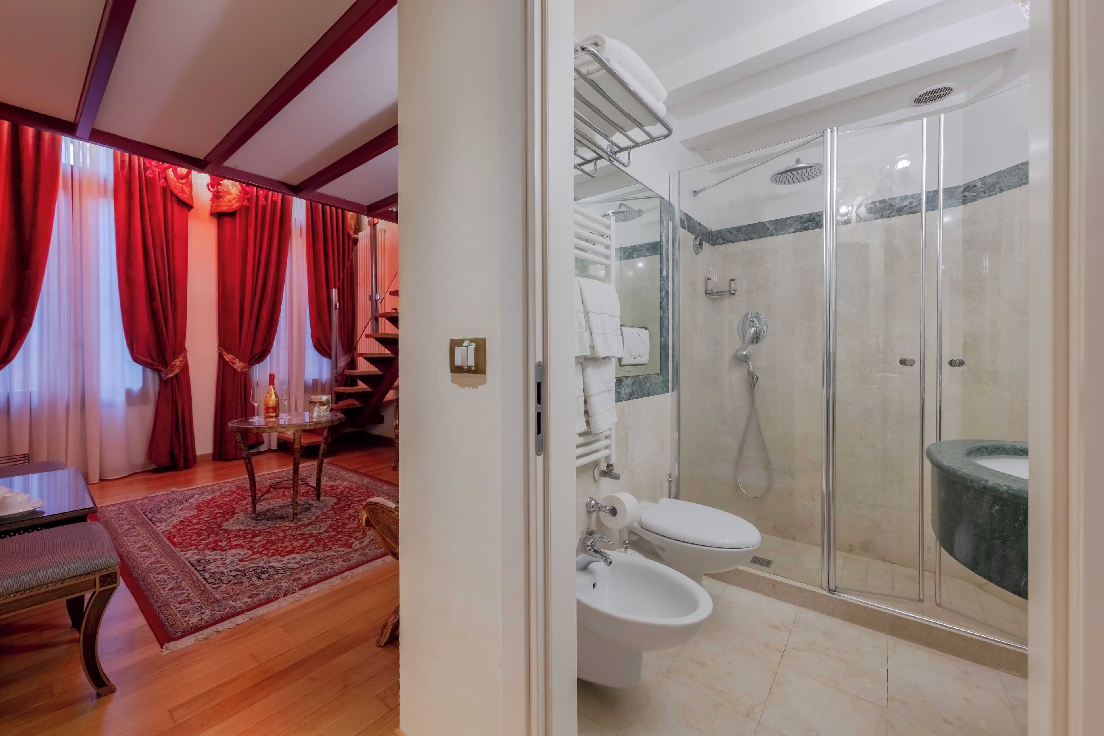 Habitación de hotel con piso de madera, alfombra roja y blanca estampada, una mesa con una silla, y zona de ducha con vidrio y lavamanos de mármol.