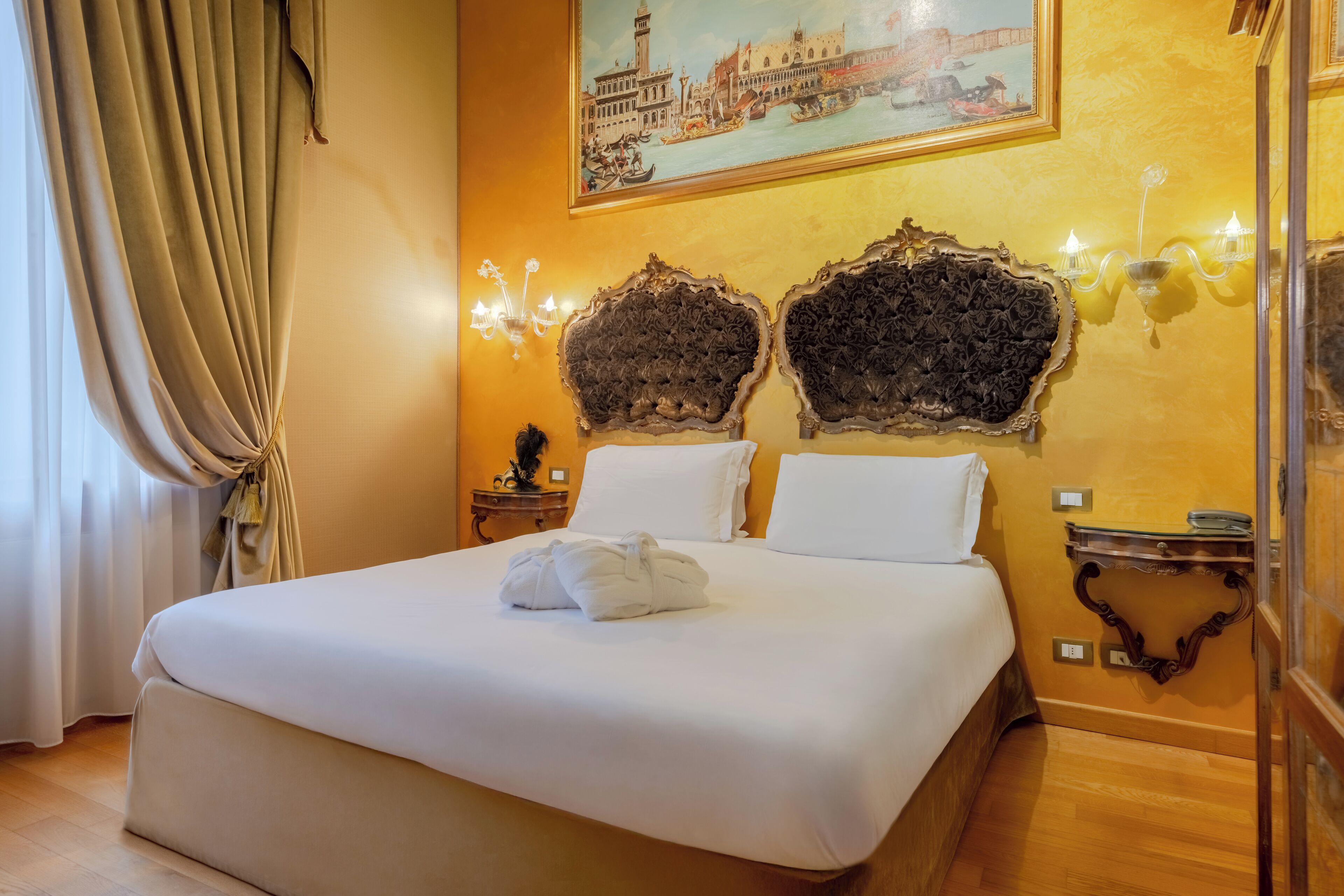 Una camera d'albergo con un letto grande, un dipinto di Venezia, applique decorate e un comodino in legno.
