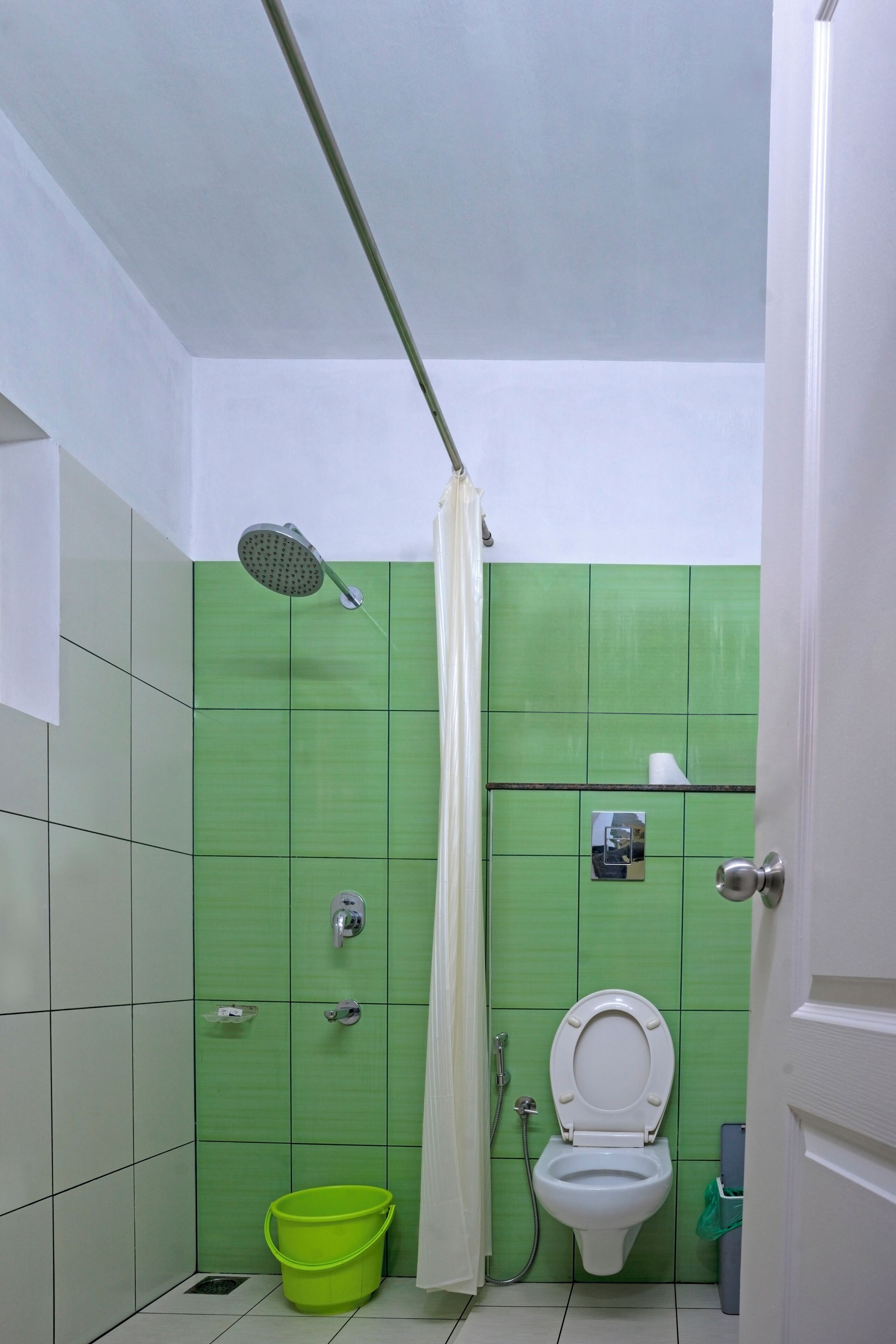 Um banheiro pequeno com azulejos verdes, vaso sanitário branco, chuveiro, um balde verde e um porta-toalhas.