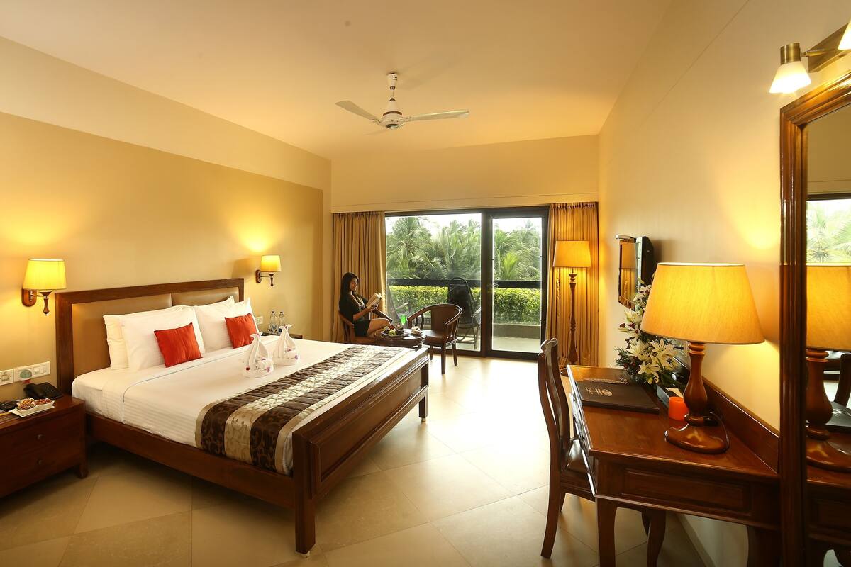 A hotel room with a bed, bedside lamps, a desk with a chair, a large mirror, and a view of the outdoors.