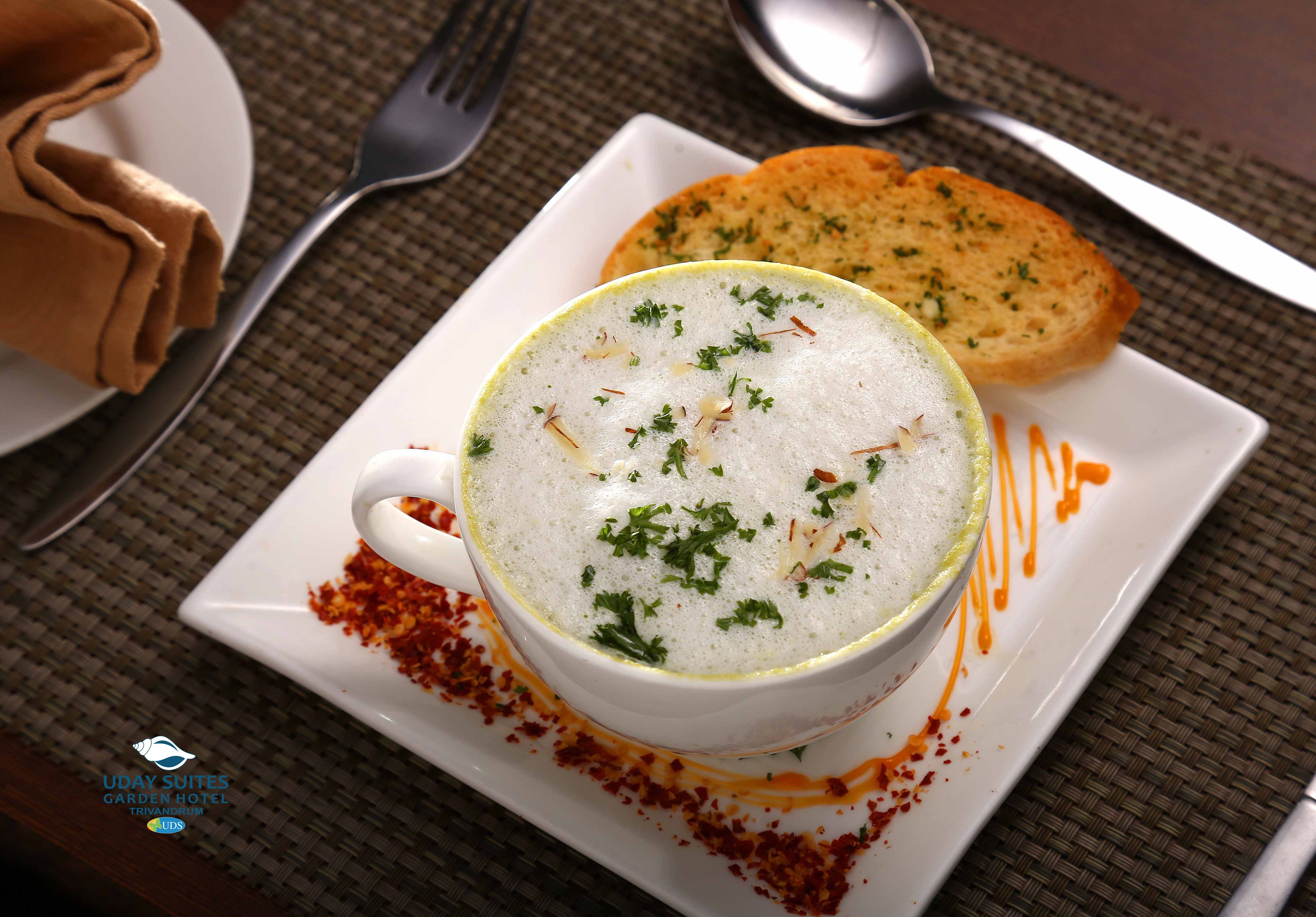 Un tazón blanco de sopa decorado con hierbas, acompañado de un trozo de pan y una cuchara sobre un plato cuadrado.
