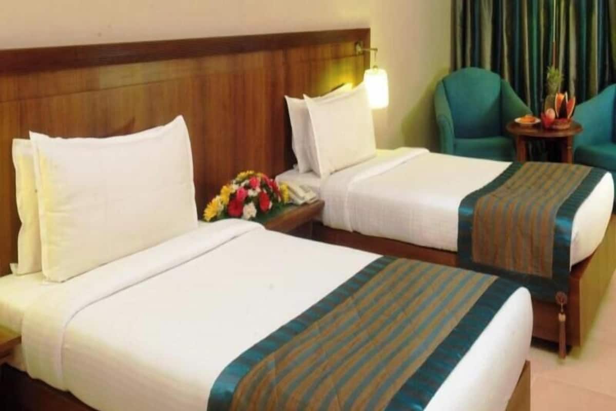 A hotel room with two beds, a wooden headboard, a nightstand with a phone and flowers, and a teal armchair.