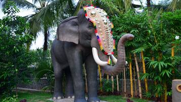 A statue of an elephant with decorative headgear, set in a garden with bamboo and palm trees.