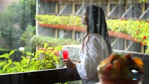 A person holding a glass of red drink, standing on a balcony with greenery and a view of a building with red accents. - Uday Suites (Thiruvananthapuram)