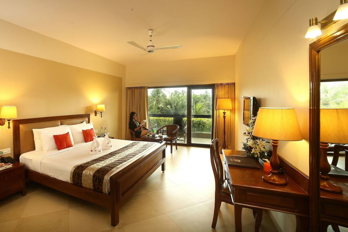 A hotel room with a bed, bedside lamps, a desk with a chair, a ceiling fan, and a view of the outdoors.